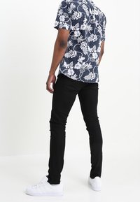 Kortärmad marinblå skjorta med vitt blommönster, matchad med åtsittande svarta byxor och vita sneakers, sedd bakifrån.