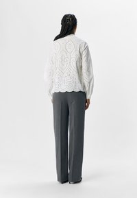 Blouse blanche brodée avec des motifs découpés, associée à un pantalon gris ajusté. La blouse possède des manches longues et un ourlet festonné.