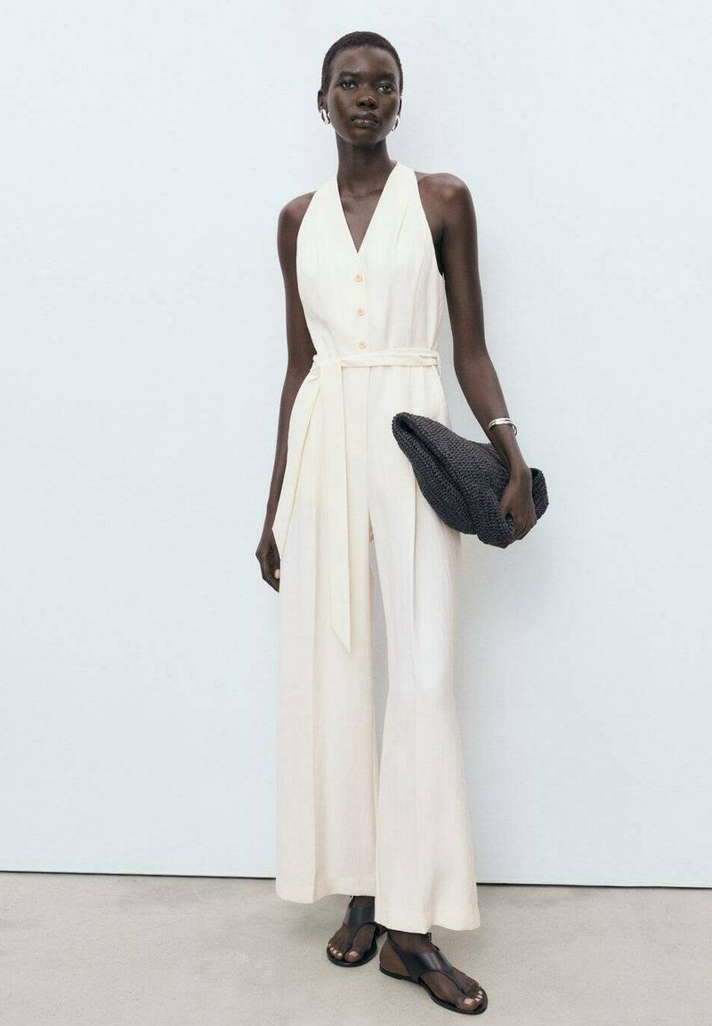 Combinaison sans manches de couleur crème avec un col en V, boutons sur le devant, taille nouée et jambes larges, associée à une pochette texturée noire et des sandales.