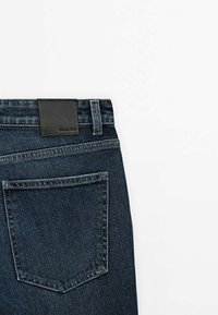 Donkerblauwe denim jeans met een gladde textuur, voorzien van een zwart leren taillebandpatch en een enkele achterzak met stikdetails.