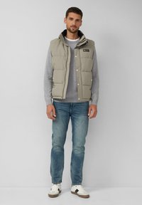 Gepolsterte Weste in Hellgrau mit hohem Kragen, Druckknöpfen und einem Logo-Patch, über einem grauen Pullover getragen und kombiniert mit blauen Jeans und weißen Sneakers.