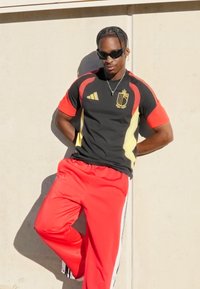 Sportieve outfit met een zwart shirt met rode mouwen en gele accenten, gecombineerd met een rode broek. Inclusief Adidas-logo en een hangende ketting.
