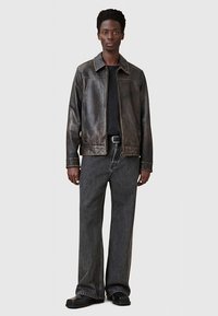 Rjava usnjena jakna z zadrgo, dvema stranskima žepoma in ravnim krojem, kombinirana z ohlapnimi sivimi kavbojkami in črnimi čevlji.