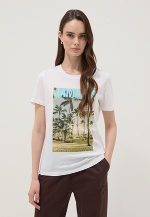 Joven mujer con cabello castaño largo que lleva una camiseta blanca con una escena de playa en Jamaica con palmeras y pantalones marrones.