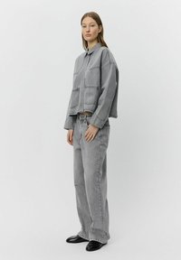 Giacca di denim grigia con tasche sul petto, abbinata a jeans denim grigi a vestibilità ampia. Il modello indossa scarpe nere. Sfondo bianco semplice.