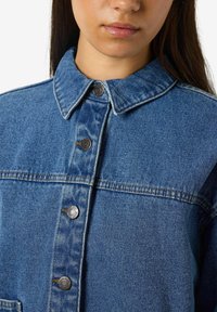 Chaqueta de denim con cuello puntiagudo, botones a presión plateados y un lavado azul clásico con costuras visibles y textura.