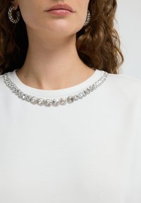 Top blanco con un escote redondo adornado con una fila de embellishments de piedras preciosas claras. La modelo lleva pendientes de aro a juego.