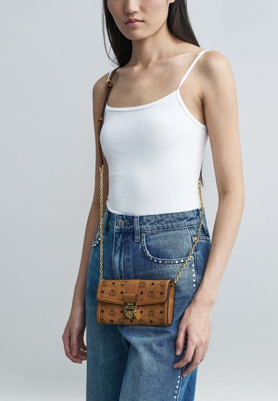 Noor naine kannab valget särkide tanki ja nupukate siniste teksadega, hoides käes väikest pruuni nahast ristiõlal kotti kuldse ahelaga.