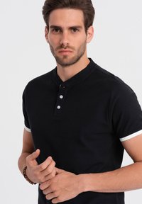 Camisa polo negra con tejido texturizado, mangas cortas con ribete blanco y tapeta de tres botones. Las manos del modelo están en la cintura.