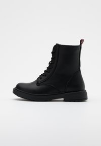Bottines en cuir noir avec un laçage, bout rond, semelle texturée et une tirette rouge à l'arrière.