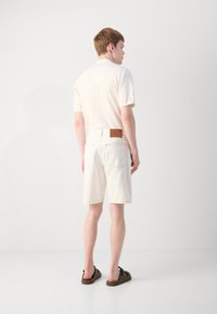 Chemise polo à manches courtes de couleur claire, shorts blancs et sandales foncées. Les shorts ont un accent en cuir au niveau de la ceinture. Design simple et moderne.