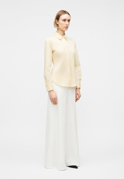 Γυναίκα όρθια, φορεμένη με ανοιχτό κίτρινο πουκάμισο με κουμπιά και μακριά μανίκια, καθώς και φαρδιά λευκά παντελόνια, μπροστά σε λευκό απλό φόντο.