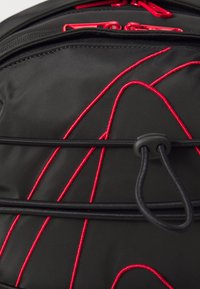 Sac à dos noir avec des motifs de couture rouge et des accessoires. Dispose de bretelles réglables et d'une surface texturée. Compartiments zippés visibles.