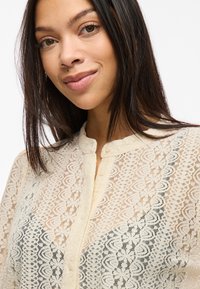 Ung kvinde med glat mørkt hår iført en cremefarvet blondebluse med knapper og blomsterprint, smilende let mod en hvid baggrund.