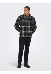 Homme debout les mains dans les poches, portant une veste à carreaux noire et grise, un pantalon noir et des bottes noires sur un fond clair uni.