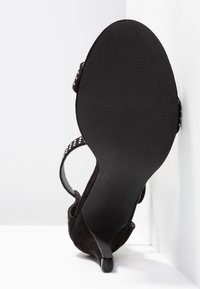 Schwarze Wildleder-Sandaletten mit Absatz, spitzer Zehenform, Knöchelriemen und einer strukturierten Gummisohle. Auffällige Aussparungen am Riemen.