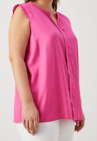 Růžová bezrukávová blůza s knoflíkovou přední částí, zdobená krajkovým lemem na ramenech, vyrobená z hladké tkaniny, spárovaná s bílými kalhotami.