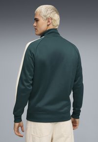 Dunkelgrüner Zip-Hoodie mit hohem Kragen, weißen Seitstreifen und gerippten Bündchen. Glatte Stoffstruktur, lockere Passform und minimalistische Markenkennung.