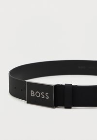 Zwarte riem met een gladde, matte textuur, voorzien van een zilveren rechthoekige gesp die het woord "BOSS" in reliëf heeft. Verstelbaar met meerdere gaatjes.