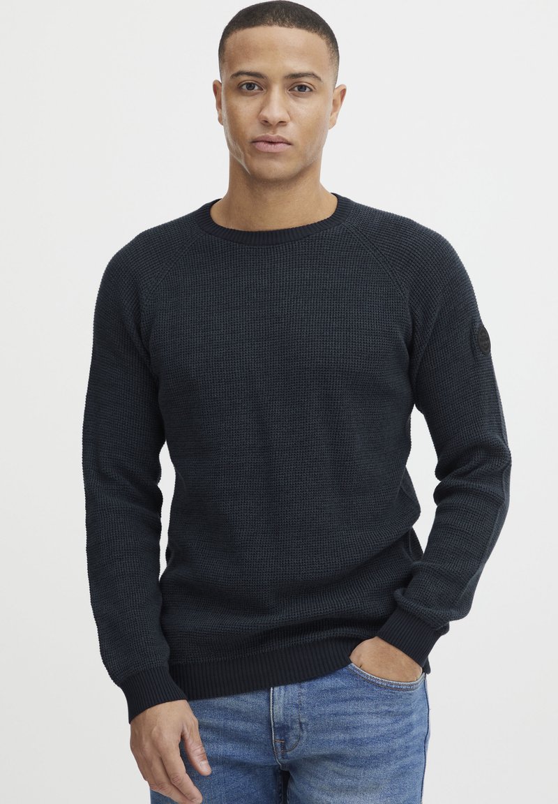 Mørk teksturert genser med ribbestrikket rund hals, lange ermer og tettsittende nederkant. Har et rundt merke på venstre erme. Brukt med blå jeans.