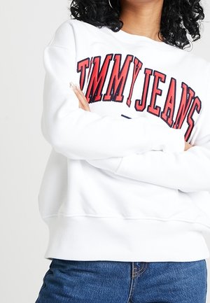 Osoba nosząca białą bluzę z napisem "TOMMY JEANS" dużymi czerwonymi literami, z założonymi ramionami, w połączeniu z niebieskimi dżinsami.