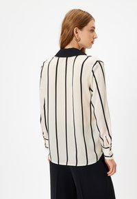 Blusa de manga larga en crema con rayas verticales negras. Presenta un cuello negro, puños abotonados y una tela suave y ligera.