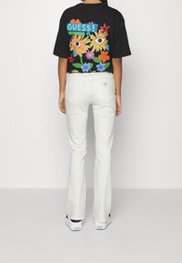 Camiseta gráfica negra con un diseño floral colorido y texto "GUESS!", combinada con jeans flare blancos y zapatillas negras. Vista trasera.