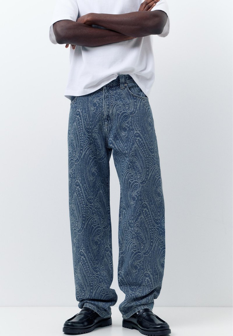Personne à la peau foncée portant un t-shirt blanc, un jean bleu ample à motif paisley et des mocassins noirs, debout les bras croisés.
