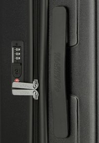 Zwarte harde koffer met een combinatie slot, dubbele ritsen en een gestructureerd oppervlak met een handvat waarop "American Tourister" is gegraveerd.