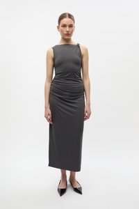 Femeie în picioare purtând o rochie midi închisă la culoare gri, fără mâneci, cu pliuri, și balerini negri cu vârf ascuțit, pe un fundal alb.
