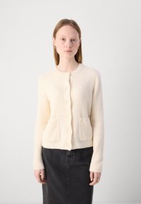 Krämfärgad texturerad cardigan med rund hals, knäppning framtill och två framfickor. Bärs över en mörk kjol.