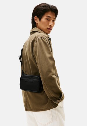 Bolso cruzado negro con textura suave, bolsillo con cremallera y correa ajustable, llevado sobre una chaqueta marrón claro y pantalones blancos.