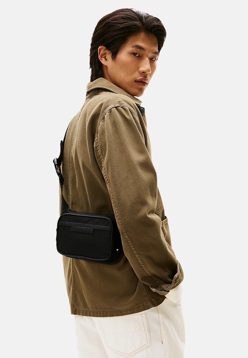 Schwarze Umhängetasche mit glatter Textur, Reißverschlusstasche und verstellbarem Riemen, getragen zu einer hellbraunen Jacke und weißen Hosen.