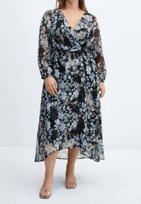 Largo vestido negro con estampado floral, con escote en V, mangas largas transparentes y cintura ceñida. Flores azules y beiges sobre un fondo negro, con un dobladillo fluido.