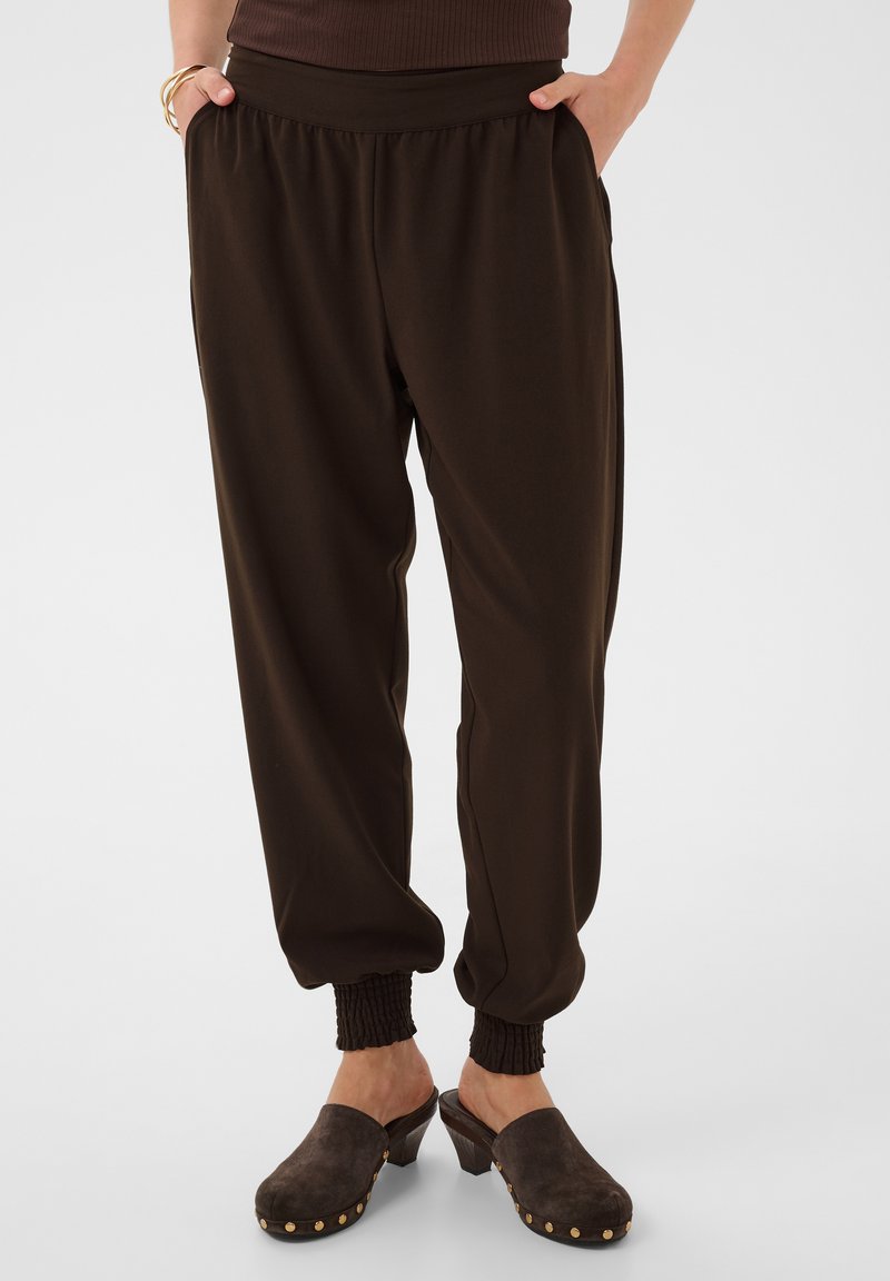 Personne portant un pantalon marron foncé avec poignets élastiques, mains dans les poches, associé à des sabots marron à talons avec des clous dorés.