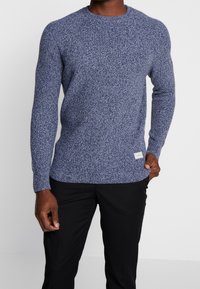 Pull en maille bleu foncé avec un motif texturé, col rond, manches longues, et une petite étiquette de marque en bas à droite. Porté avec un pantalon noir.