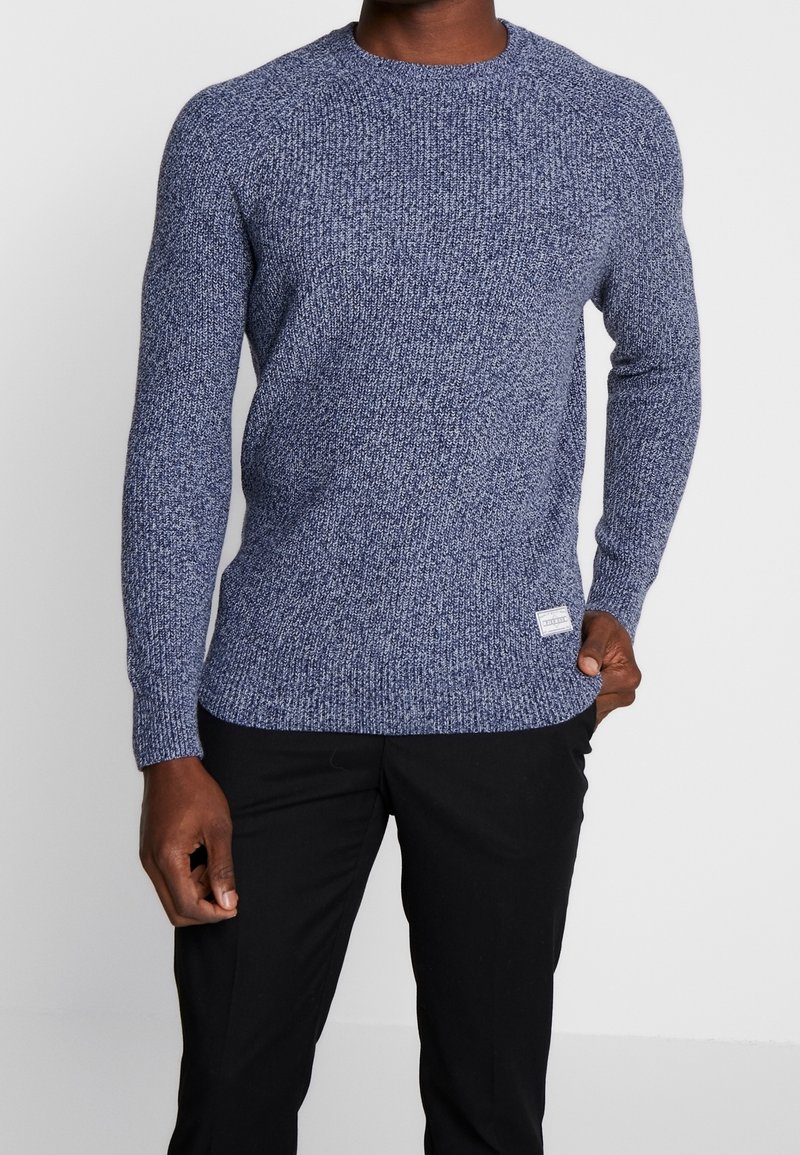 Pull en maille bleu foncé avec un motif texturé, col rond, manches longues, et une petite étiquette de marque en bas à droite. Porté avec un pantalon noir.