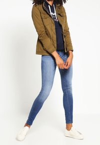 Olivgrüne Militärjacke mit zwei Brusttaschen über einem navyblauen Hoodie, kombiniert mit hellblauen Skinny Jeans und weißen Sneakers.