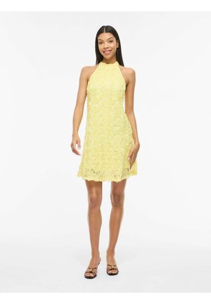 Jauna moteris šypsosi, dėvi geltoną tekstūruotą gėlių rašto suknelę be rankovių ir juodus atvirus sandalų pirštus, stovinčią prieš paprastą baltą foną.