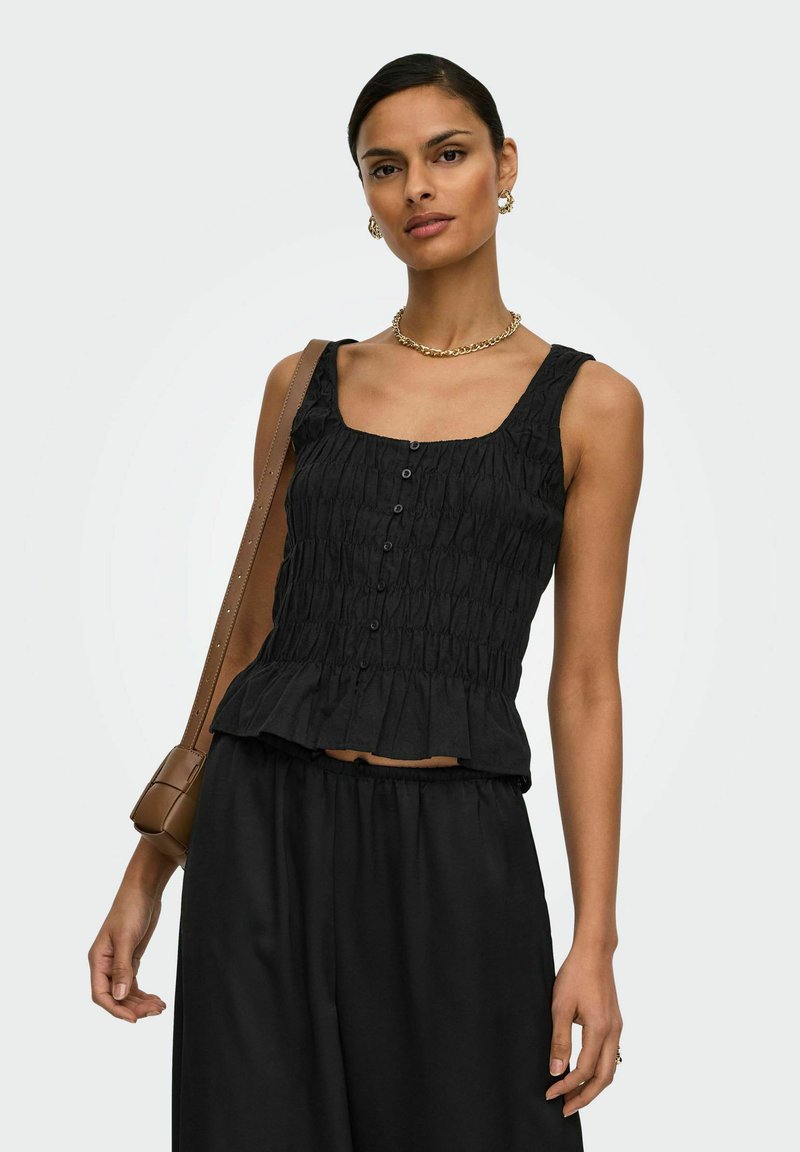 Femme portant un haut noir sans manches froncé avec des boutons, un pantalon noir, un collier chaîne doré, des boucles d'oreilles créoles, et portant un sac à bandoulière marron.