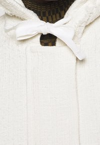 Białe teksturowane okrycie wierzchnie z kapturem, wyposażone w zawiązywaną wstążkę. Wnętrze jest wykonane z ciemnej dzianiny w paski.