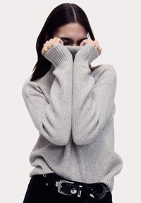 Maglione grigio chiaro a maglia con colletto alto, texture a coste e maniche lunghe; indossato con pantaloni neri con una fibbia lucida.