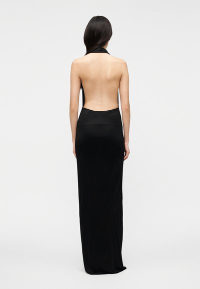 Hosszú sötét hajú nő, ujjatlan fekete, hátul kivágott estélyi ruhában, egy egyszínű világosszürke háttér előtt áll.