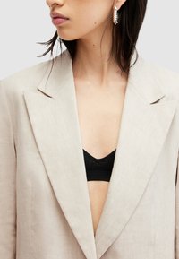 Blazer di lino beige con colletto strutturato, scollatura a V profonda e tasche; indossato sopra un reggiseno nero, in tessuto testurizzato.