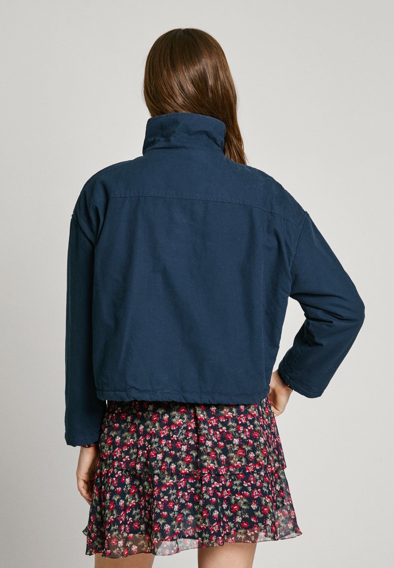 Marineblau gekürzte Jacke mit hohem Kragen und langen Ärmeln, aus robustem Stoff. Getragen über einem blumigen Rock mit Lagen.