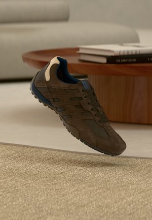 Brown athletic sneaker with black and beige details hovering above a textured beige carpet, with a round wooden table and books in the background.