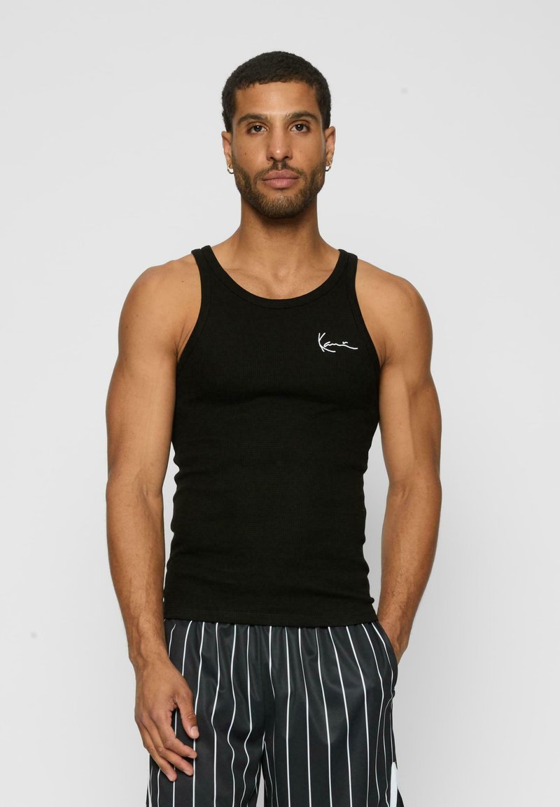 Sort ribbet tanktop med rund halsudskæring og hvidt logo accent på venstre bryst, bæres med grå stribede shorts. Glat stofstruktur.
