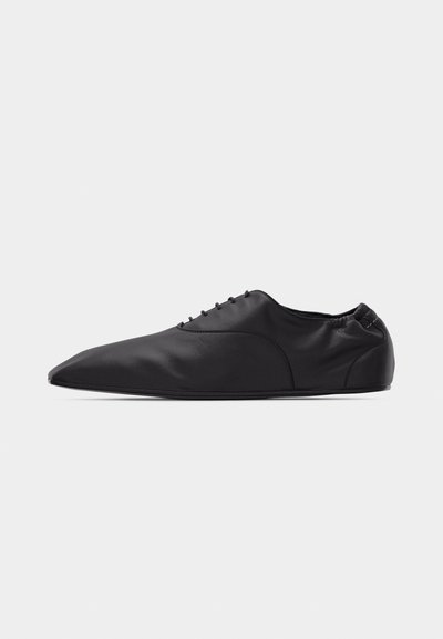 Zapato de cuero negro con una punta afiladamente puntiaguda, cordones delanteros y talón trasero elástico, mostrado de perfil sobre un fondo blanco.