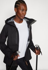 Veste imperméable noire avec un intérieur gris, poches zippées et capuche ajustable. Une chemise blanche en dessous et des bâtons de randonnée en main.