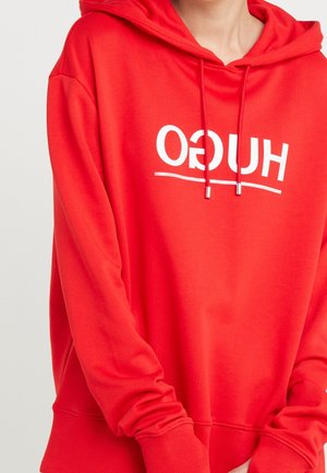 Personne portant un sweat à capuche rouge avec du texte blanc inversé "HUGO" et des cordons, montrant la partie supérieure du torse et les bras croisés.